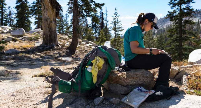 A person sits on a rock while their backpack rests on the ground 